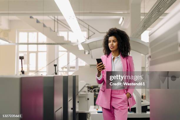 female professional with mobile phone looking away while standing amidst machines in industry - pant suit stock-fotos und bilder