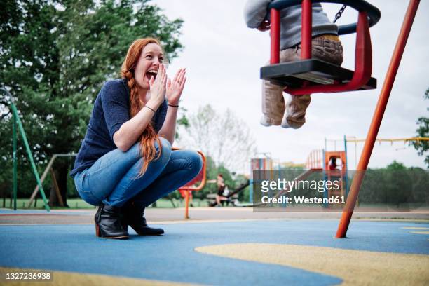 happy mother clapping while crouching near son sitting on swing in playground - britain playgrounds fotografías e imágenes de stock