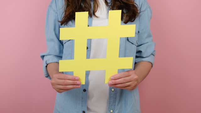 Closeup of young girl demonstrate large big yellow hashtag sign, concept of trendy social media posts and blogging, viral web content, internet promotion, isolated over pink studio background wall
