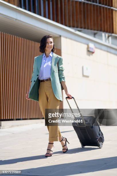 smiling businesswoman walking with suitcase on footpath - business woman suitcase stockfoto's en -beelden