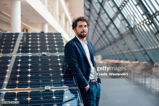 businessman with hands in pockets leaning on solar panel - zonnecellen stockfoto's en -beelden