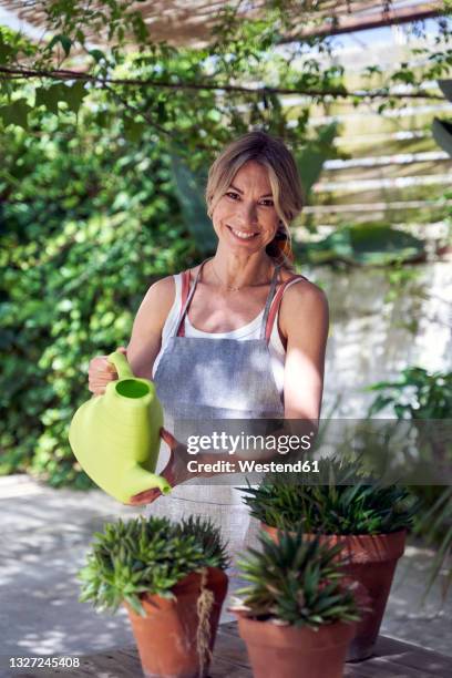 smiling mature woman watering plants while standing in garden - lush backyard stock pictures, royalty-free photos & images