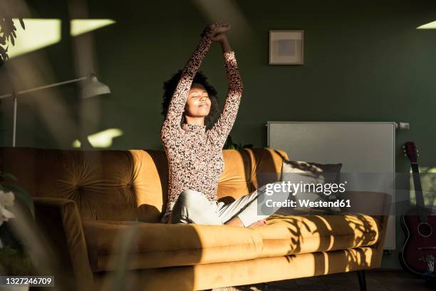 woman with hands raised sitting on sofa at home - relajación fotografías e imágenes de stock