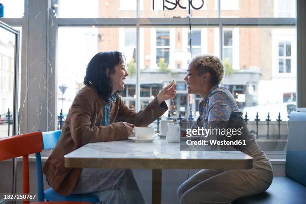 happy young woman feeding food to girlfriend sitting in cafe - lesbian date stock pictures, royalty-free photos & images