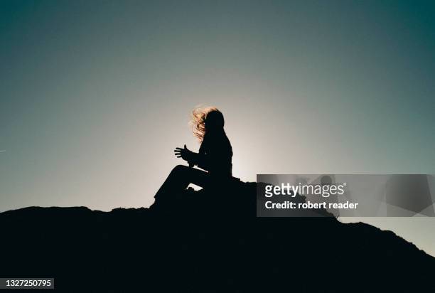 silhouette of a person sitting on cliff with hair blowing in the wind - hair wind stock pictures, royalty-free photos & images