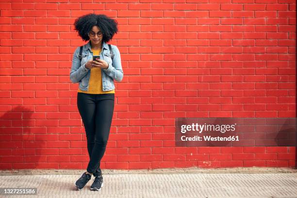 young woman using smart phone while standing in front of red brick wall - black woman standing stock-fotos und bilder