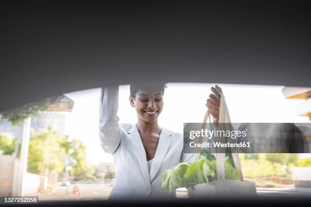 smiling businesswoman closing car trunk while holding bag of vegetables - closing car boot fotografías e imágenes de stock