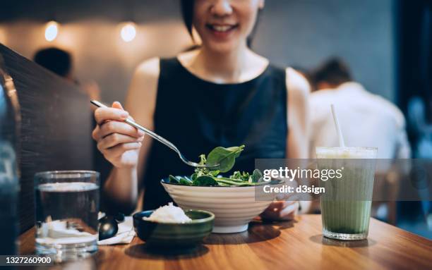 cropped shot of smiling young asian woman eating fresh vegan salad with a glass of green smoothie in a restaurant, enjoying a healthy meal. green living. healthy eating lifestyle. vegetarian and vegan diet concept - dinner business stock-fotos und bilder