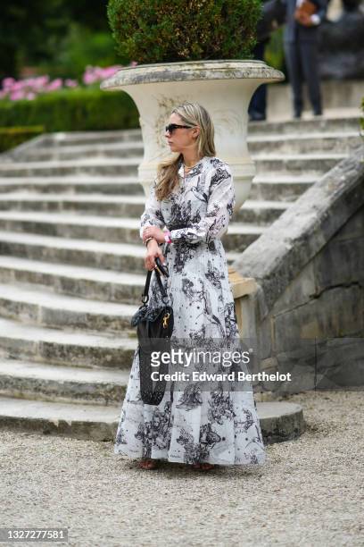 Guest wears black sunglasses, a long sleeves white with black print patter ruffled long flowing dress from Dior, gold rings, a silver watch, a black...