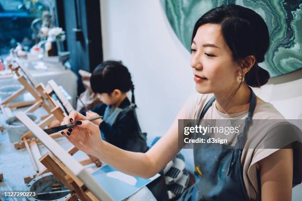 young asian mother and little daughter having a painting class together. painting on a canvas with paintbrush and colourful acrylic paints. having a relaxing afternoon - desenhar atividade - fotografias e filmes do acervo