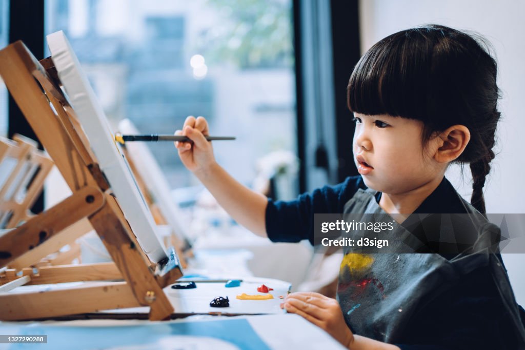 Concentrated little Asian girl painting on a canvas with paintbrush and colourful acrylic paints in a painting class