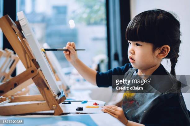 concentrated little asian girl painting on a canvas with paintbrush and colourful acrylic paints in a painting class - drawing activity stock pictures, royalty-free photos & images