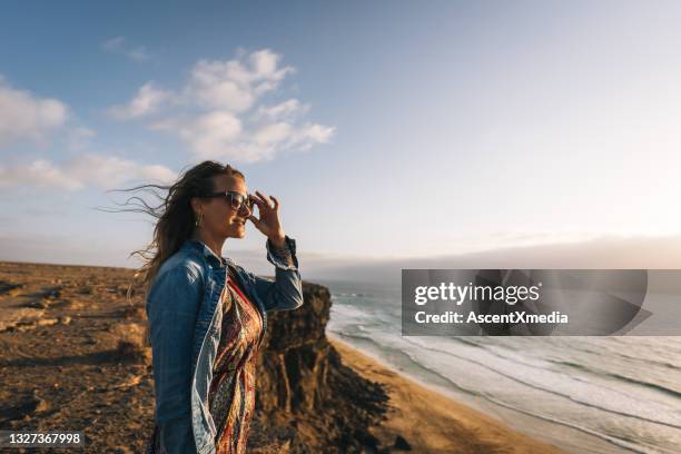 young woman standing on sunny coastal cliff puts on sunglasses - lanzarote stock pictures, royalty-free photos & images