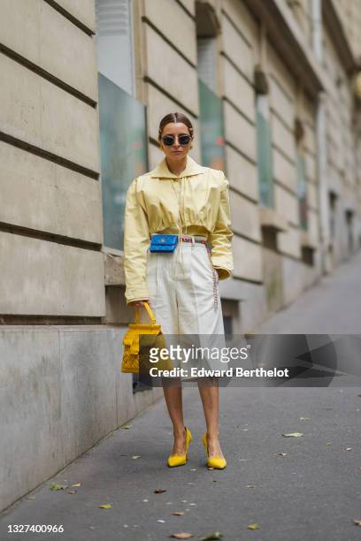 Julia Comil wears sunglasses, gold hair clips, gold earrings, a pale yellow shirt, a matching pale yellow adjustable hoodie vest / short jacket, a...