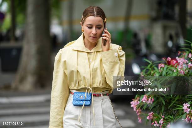 Julia Comil wears sunglasses, gold hair clips, gold earrings, a pale yellow shirt, a matching pale yellow adjustable hoodie vest / short jacket, a...