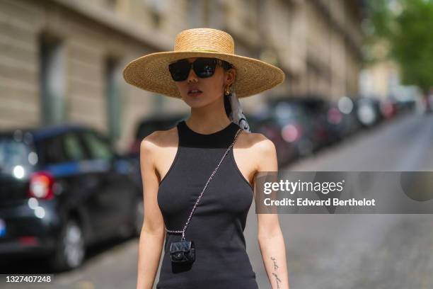 Xiayan wears sunglasses, a white with black print pattern scarf in the hair, gold earrings, a beige wicker hat with a green ribbon, a black...