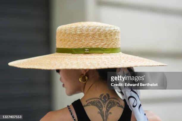 Xiayan wears sunglasses, a white with black print pattern scarf in the hair, gold earrings, a beige wicker hat with a green ribbon from Chanel, a...