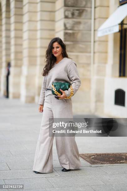 Gili Biegun wears gold earrings, a gold chain necklace, a gray V-neck long sleeves pullover, matching gray large pants, a black shiny leather belt...