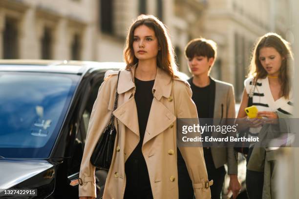 Model wears a black t-shirt, a beige long trench coat from Burberry, black denim flared jeans pants, a black shiny leather shoulder bag, outside...