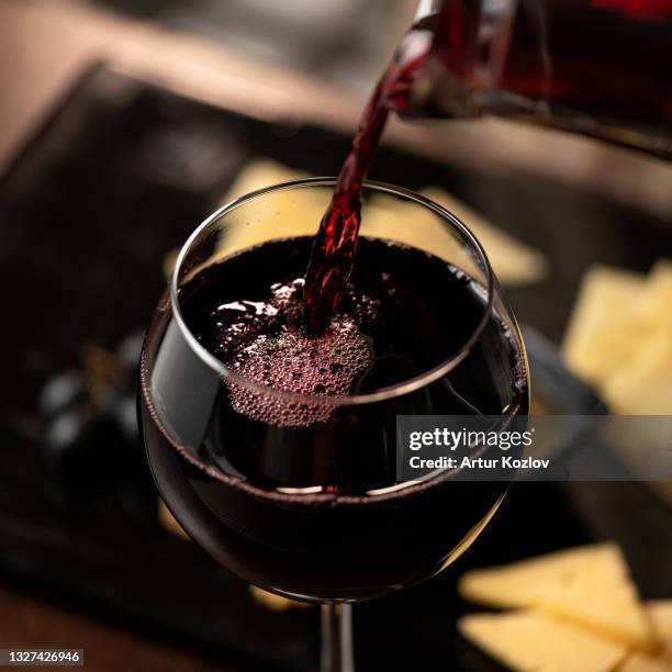 expensive red wine pouring into wine glass. wineglass with merlot or sauvignon on blurred background. close up shot. luxury alcoholic drink or beverage - maroon fotografías e imágenes de stock