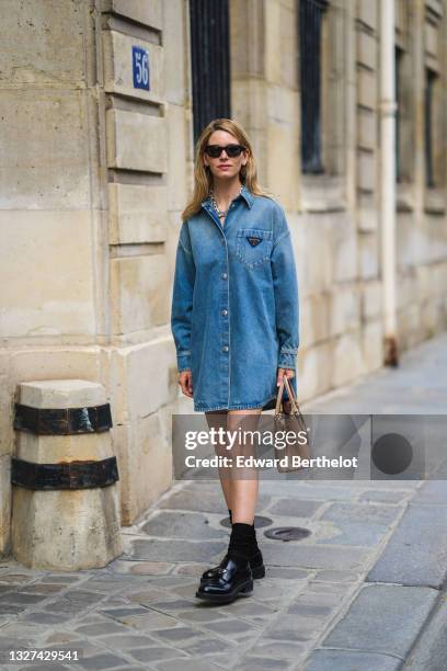 Natalia Verza wears gold earrings, black sunglasses, a blue denim Prada shirt dress, a silver large chain necklace, a brown grained leather Prada...