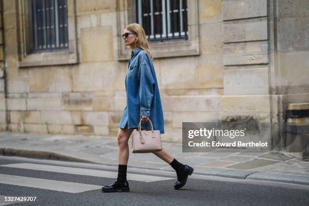 Natalia Verza wears gold earrings, black sunglasses, a blue denim Prada shirt dress, a silver large chain necklace, a brown grained leather Prada...