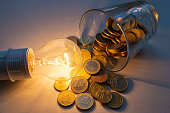 Light bulb lit with glass full of coins next to it.