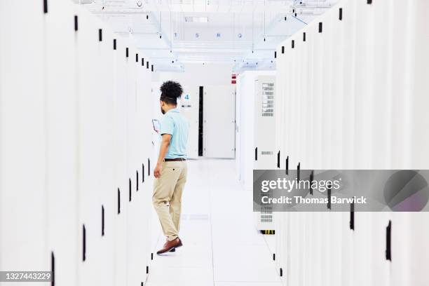 wide shot of male it professional looking at smart phone while walking through server racks in data center - pantalon beige photos et images de collection