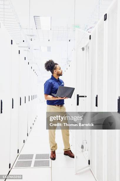 wide shot of male computer engineer using laptop while working on server in data center - pantalon beige photos et images de collection