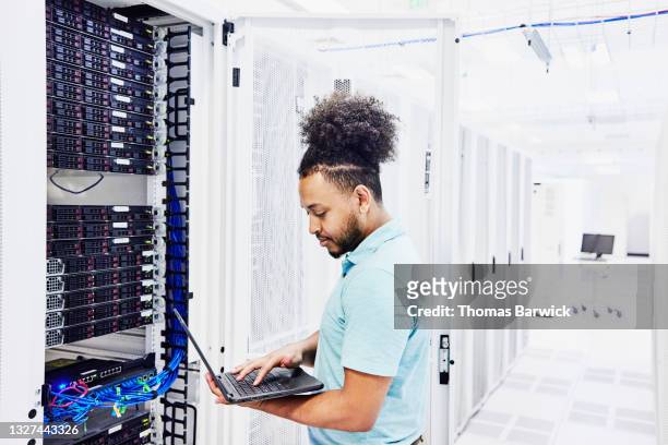 medium shot of male it professional looking at laptop while working on server in data center - cloud computing stock-fotos und bilder