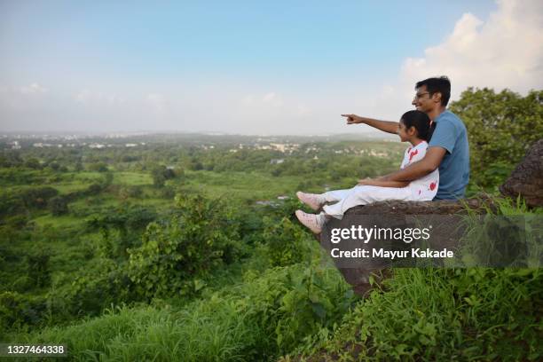 father and daughter sitting on a rock edge on a cliff in the forest - enjoy monsoon stock-fotos und bilder