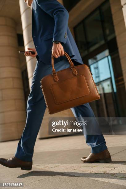 una vista de cerca de un hombre con un traje clásico azul que pasa por el centro de negocios, llevando una bolsa marrón para computadora portátil en una mano y un teléfono en otra. - maletín para portátil fotografías e imágenes de stock