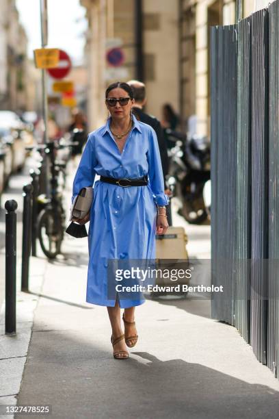 Guest wears black sunglasses, a gold sunglasses chain, a gold chain necklace, a blue long oversized shirt dress, a black leather belt, a silver...