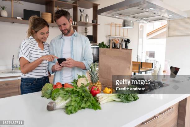 coppia che fa la spesa online utilizzando un'applicazione mobile - couple in kitchen foto e immagini stock