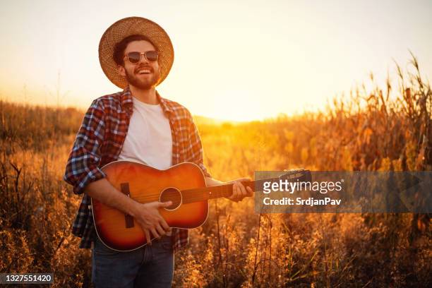 fröhlicher, hübscher mann, der während des sonnenuntergangs im freien gitarre spielt - country and western music stock-fotos und bilder