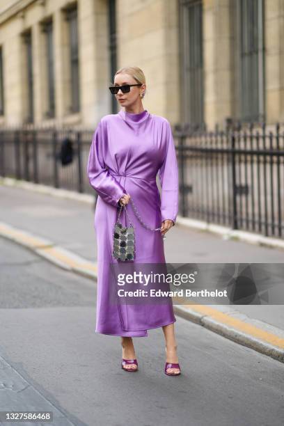 Justyna Czerniak wears black sunglasses, gold earrings, a shiny purple turtleneck long sleeves flowing long dress, silver yoke handbag, purple shiny...