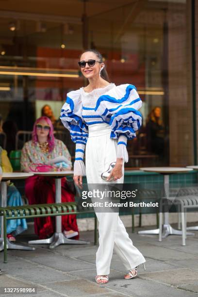 Guest wears a silver and crystals pendant large earring, black sunglasses, a white with blue striped ruffled large puffy long sleeves t-shirt, white...