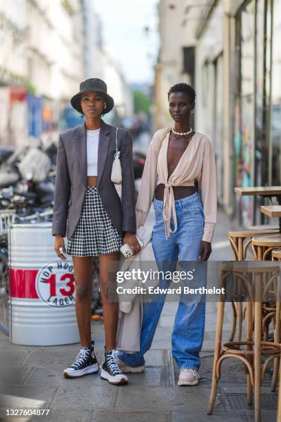Model wears a black bob hat, a white ribbed t-shirt, gold chain pendant necklaces, a gray blazer jacket, a black and white checkered pattern pleated...