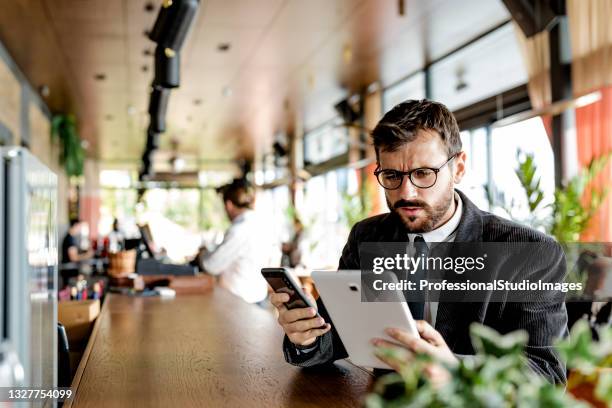 un giovane e felice uomo d'affari sta digitando messaggi sul suo telefono cellulare e tenendo in mano un tablet digitale mentre è seduto nella caffetteria moderna. - man suit using phone tablet foto e immagini stock