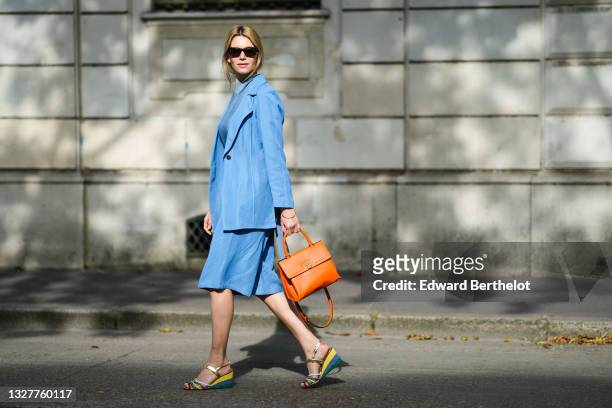Natalia Verza wears sunglasses, earrings, a blue wool pullover, a blue flowing blazer jacket, blue knees flowing short, an orange shiny leather...