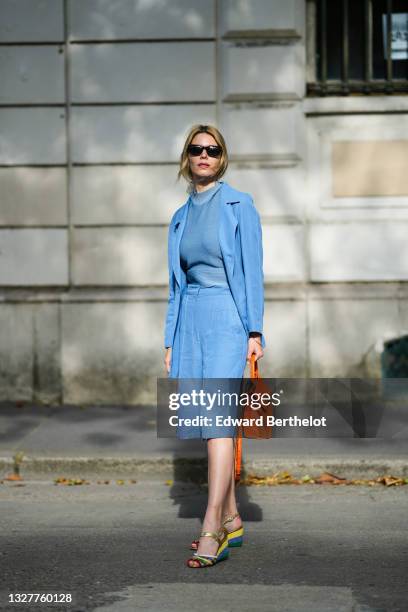 Natalia Verza wears sunglasses, earrings, a blue wool pullover, a blue flowing blazer jacket, blue knees flowing short, an orange shiny leather...
