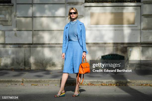 Natalia Verza wears sunglasses, earrings, a blue wool pullover, a blue flowing blazer jacket, blue knees flowing short, an orange shiny leather...