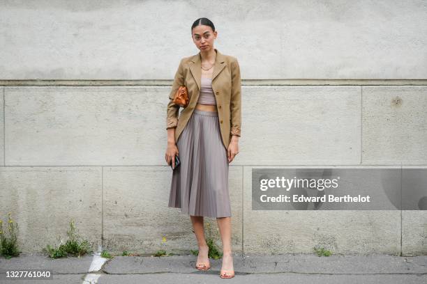 Maria Decremps wears gold necklace, a brown blazer jacket, a beige crop-top, a brown shiny leather shoulder bag, a gray pleated / accordion midi...