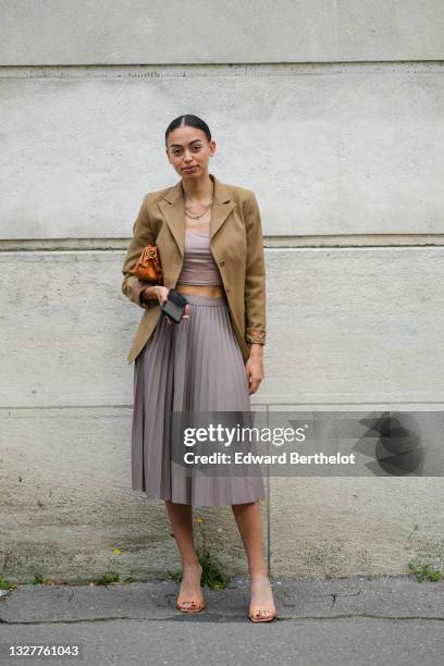 Maria Decremps wears gold necklace, a brown blazer jacket, a beige crop-top, a brown shiny leather shoulder bag, a gray pleated / accordion midi...