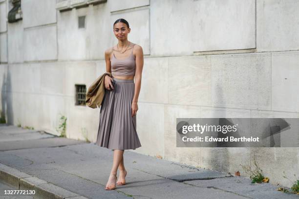 Maria Decremps wears gold earrings, gold necklaces, a brown blazer jacket, a beige crop-top, a gray pleated / accordion midi skirt, beige shiny...