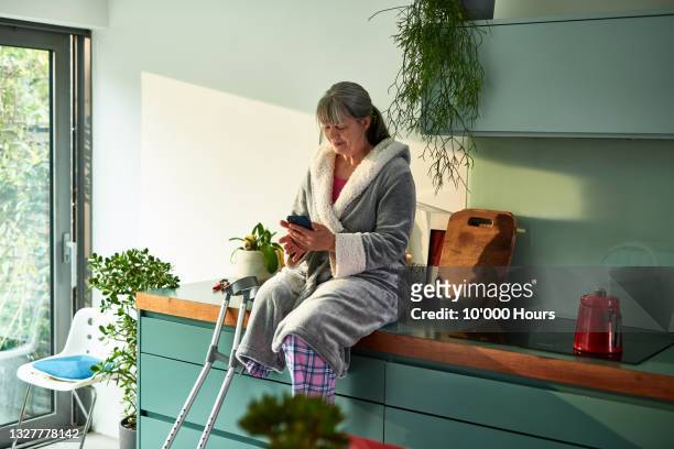 mature amputee woman sitting on kitchen bench using mobile phone - körperliche behinderung stock-fotos und bilder