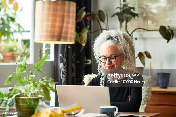 grey haired woman at home smiling in front of laptop - senior women foto e immagini stock