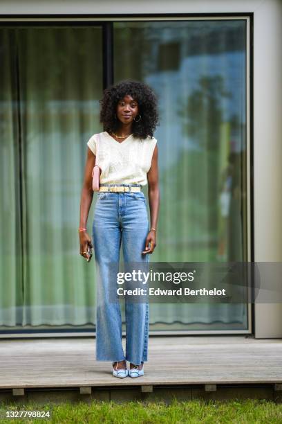 Guest wears a white knit woven wool sleeveless v-neck pullover, a golden necklace, a beige belt, blue flared denim jeans pants, blue shoes, a pastel...