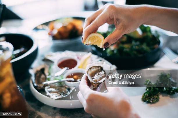 close up of a woman's hand squeezing lemon juice on to a fresh oyster, enjoying a scrumptious meal in a restaurant. eating out lifestyle - fine art stock-fotos und bilder