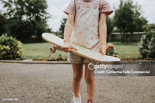 cute young girl in spotty dungarees holds a spotty skateboard - skate sports footwear stock pictures, royalty-free photos & images